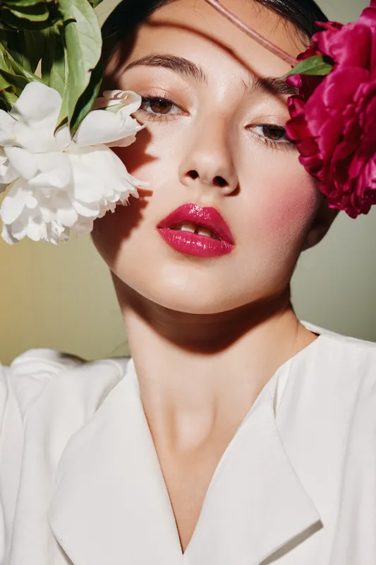 Close-up of a young woman with glossy red lips and radiant skin surrounded by vibrant peony flowers. Soft lighting enhances the beauty fashion and creative makeup concept.