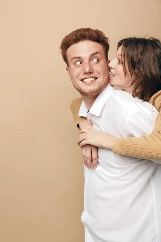 Happy young couple in casual clothes share joyful moment, woman hugging man from behind on beige background. Concept of love, friendship, and positive relationship.