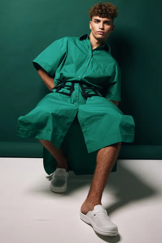 Young man with curly hair wearing green oversized shirt and shorts sitting against dark green background with white slip on shoes in studio lighting.