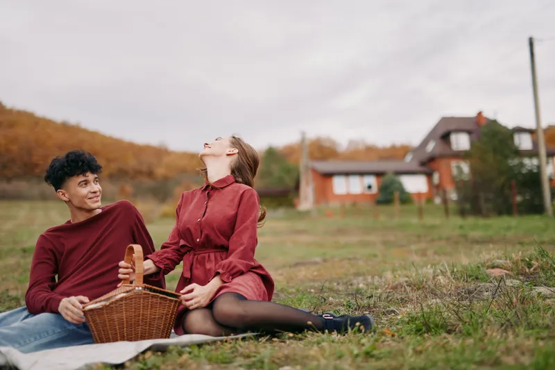 An authentic candid moment of a couple sharing a picnic in a rural field, warm tones, natural light, simple companionship, sincere smiles, quiet joy, unposed scene.