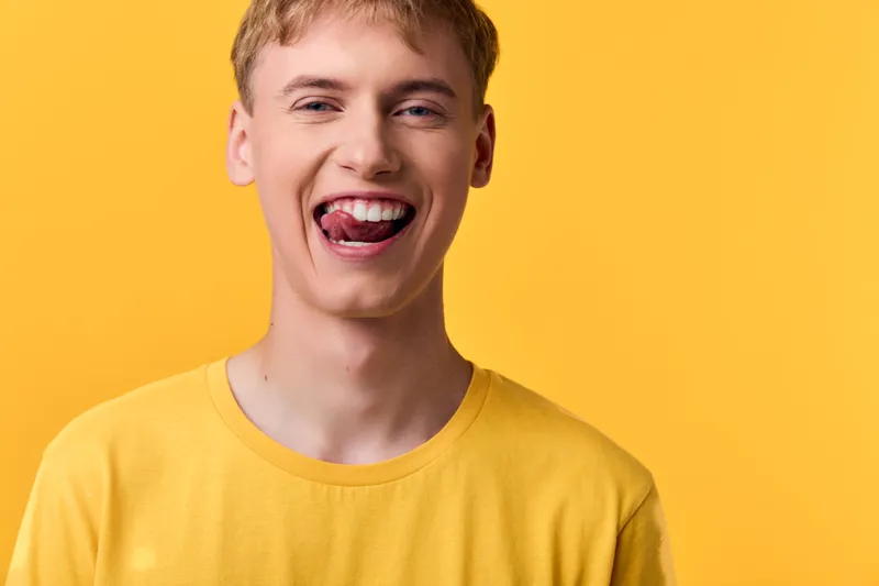 Playful, joyful, yellow background with a young man sticking out his tongue, showcasing a bright smile and expressive face, casual and fun mood suitable for lifestyle, youth culture and upbeat