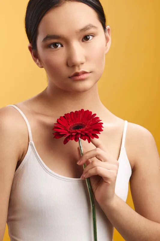 asian woman holds a red flower close to her chest against a warm yellow backdrop, conveying calm elegance and focus with a soft, contemplative expression.