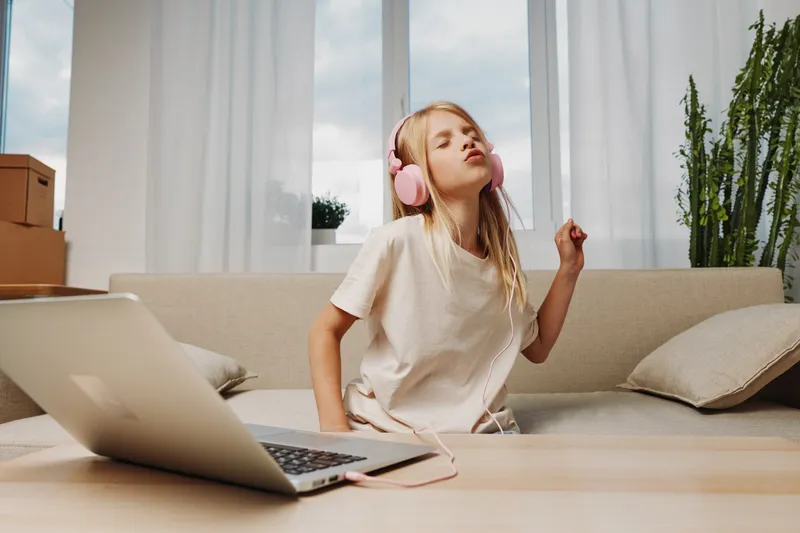 Young girl with long blonde hair wearing pink headphones enjoys music at home, sitting on a sofa with a laptop on the table. Bright natural light fills the cozy living room space.