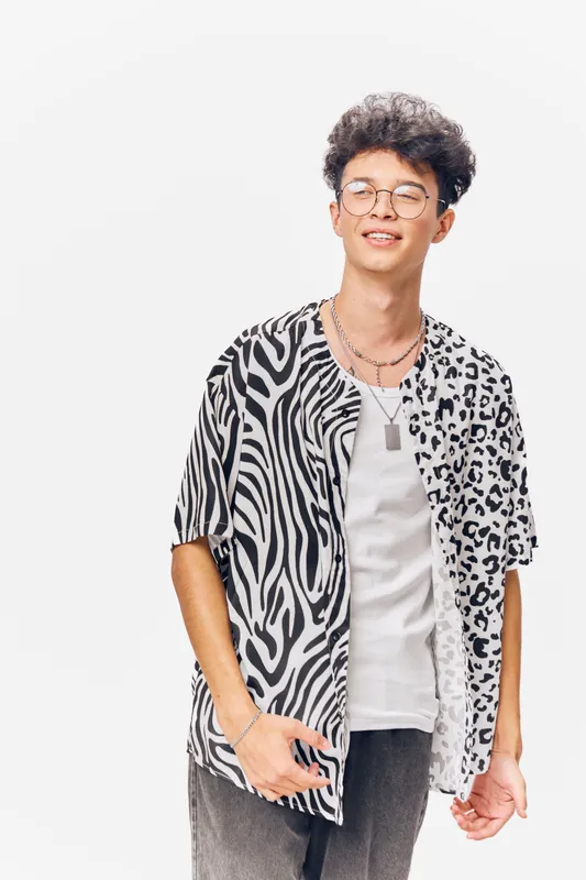 young man wearing a black and white patterned shirt with glasses smiling confidently in a studio setting with a plain white background and casual style