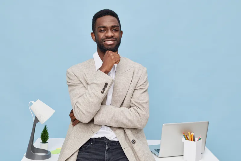 Confident smiling man in beige blazer and white shirt standing in modern office. Casual pose with hand on chin, positive expression, minimalistic workspace with laptop, lamp, plant, and stationery. Pe
