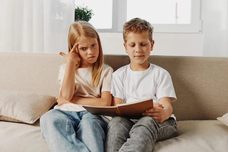 Two children sitting on a sofa reading a book together, girl looks confused while boy is focused. Concept of learning, childhood, education, and concentration at home.
