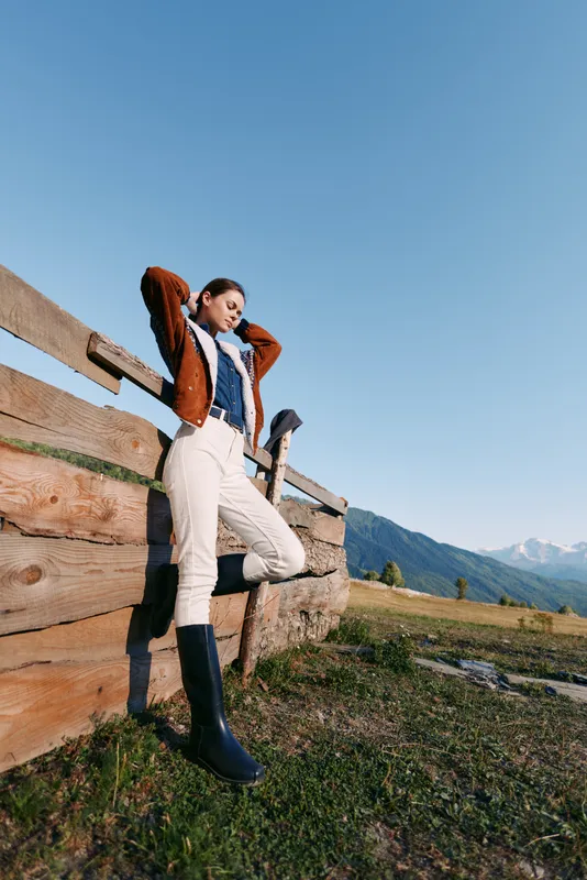 Woman by wooden fence in mountains and countryside, wearing boots and jacket in a meadow, portrait pose enjoying sunlight and serene outdoor landscape with blue sky.