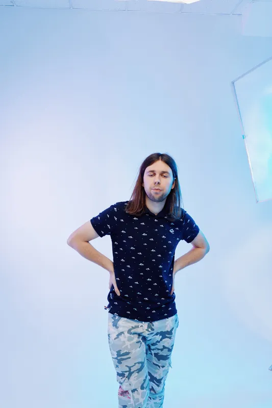 Young man with long hair wearing a dark patterned shirt and camouflage pants stands with hands on hips in a bright studio. Concept for fashion, casual style, and confidence.