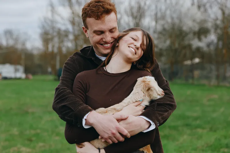 A joyful couple outdoors, embracing a small goat kid in a grassy field, wearing cozy sweaters. A candid lifestyle moment highlighting affection, companionship, and rural life.