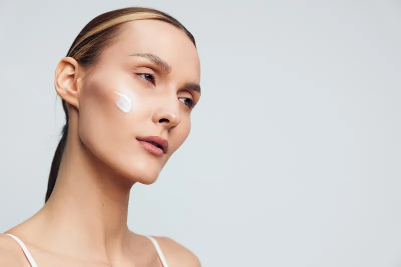 Young woman with clean skin applying cream on cheek, neutral background, natural light, skincare routine, beauty concept, calm expression, minimal makeup, closeup portrait