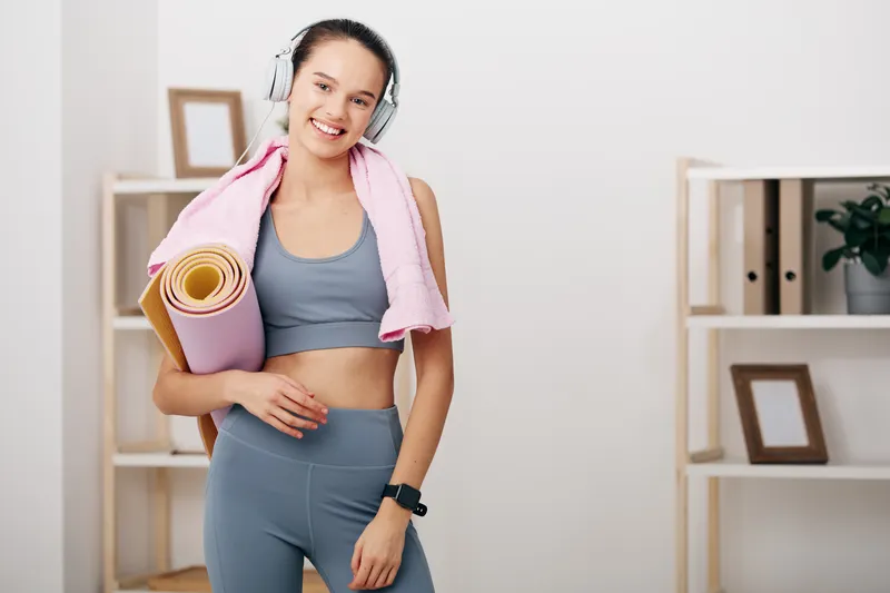 Fitness woman in a home gym, wearing headphones and a gray workout set, holding a rolled yoga mat with a towel over her shoulders, smiling after a workout near shelves.