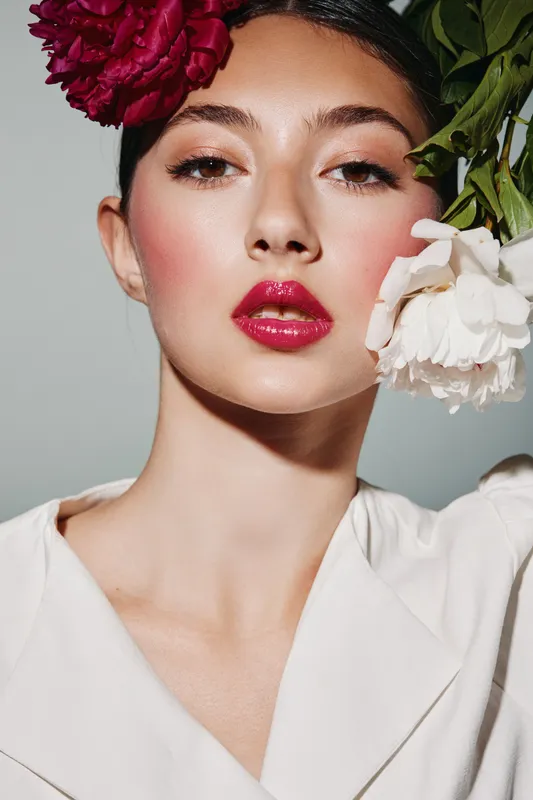 Young woman with bold red lipstick and complete makeup wears white collar with flowers on head. Her face shows elegance and beauty with flawless skin in a studio shot. Creative fashion theme.