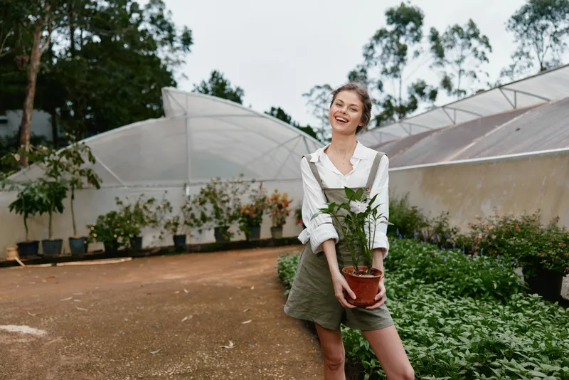 Portrait of a joyful young woman holding a potted plant in a greenhouse, showcasing sustainable gardening and eco friendly living, wearing stylish casual attire