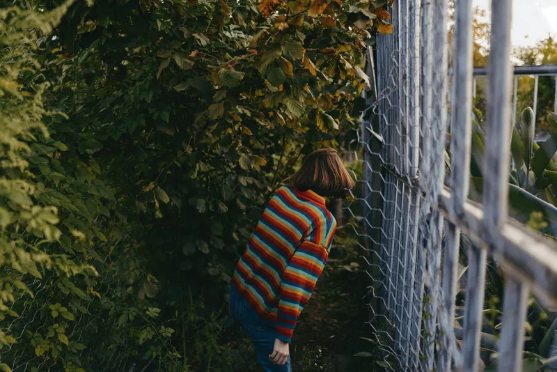Child in a striped sweater peeking through a metal fence beside overgrown bushes, outdoor curiosity and exploration, young girl discovering a hidden garden path in nature