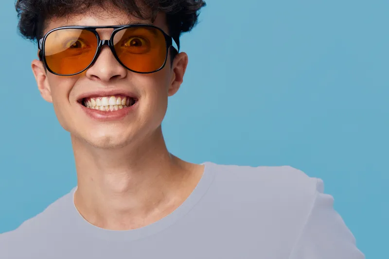 smiling man with orange sunglasses showing teeth expression against blue background, casual white t-shirt, joyful and youthful mood, studio portrait