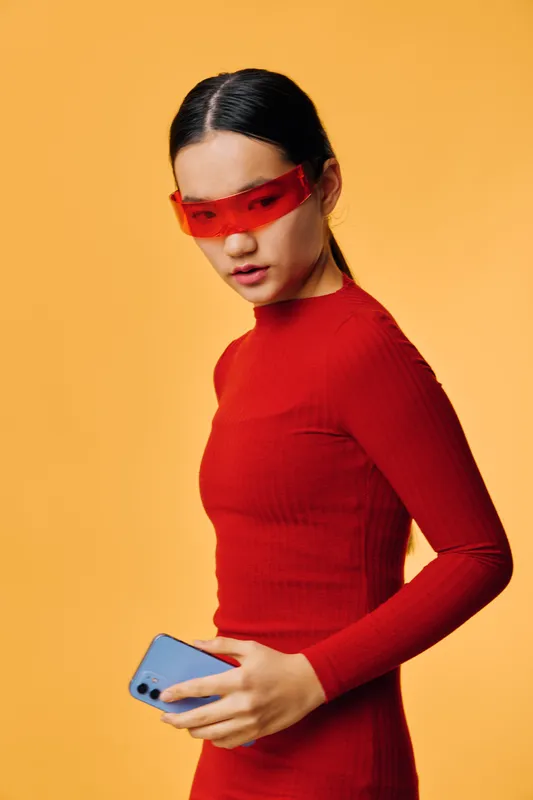 Young woman in red dress and red sunglasses holding smartphone with serious expression on orange background. Fashionable model with modern accessory and smooth hairstyle.