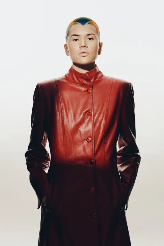 Asian woman wearing a long red leather coat with hands in pockets, short colorful hair, modern fashion style, and neutral facial expression on white background.
