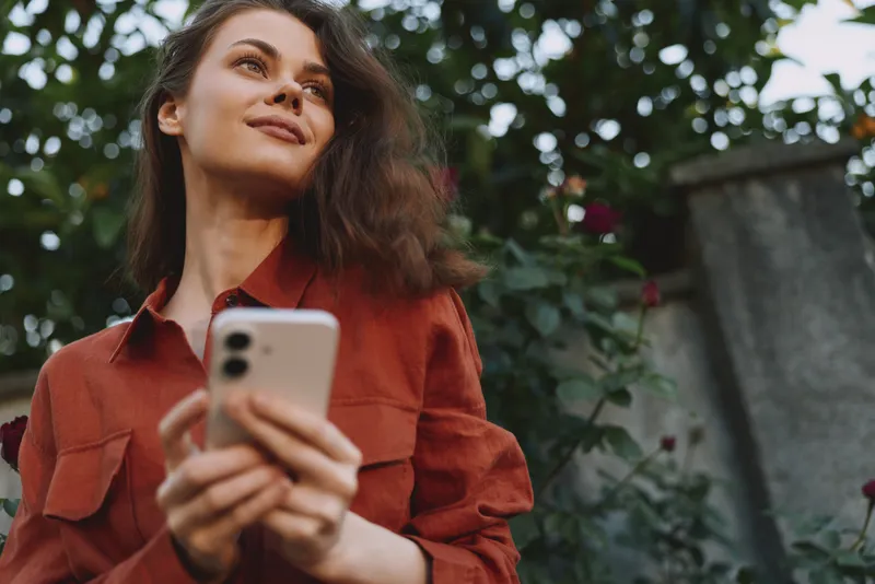 A woman outdoors in a red shirt holds a phone, smiling softly in natural light. A lifestyle portrait capturing confidence, calm, and relaxed mood in everyday moments