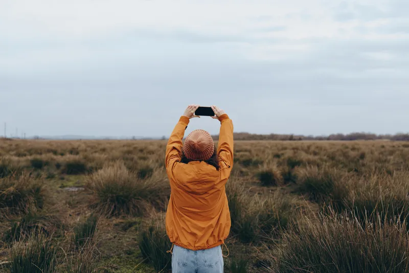Woman in orange jacket standing in grassy field outdoors, arms raised behind head, back view, serene nature lifestyle, freedom and relaxation, soft daylight, wide landscape, autumn mood