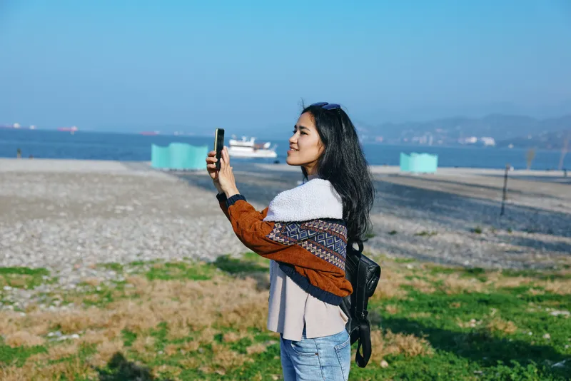 Woman on a sunny beach captures a selfie with phone, backpack and scarf, ocean and horizon in the distance, casual travel scene, bright outdoor moment, candid lifestyle photography