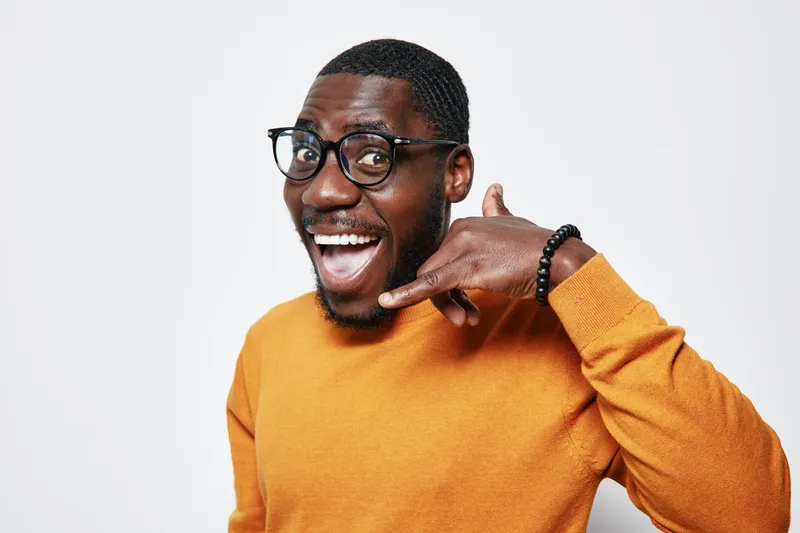 African American man with dark skin wearing glasses and orange sweater smiling and making phone call gesture with hand isolated on white background for communication concept.
