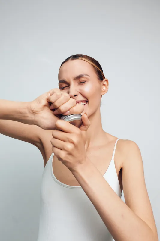 Playful woman in a white tank top pops a can with a smile, eyes closed, capturing candid humor in an indoor studio for lifestyle, wellness, and cheerful moments.