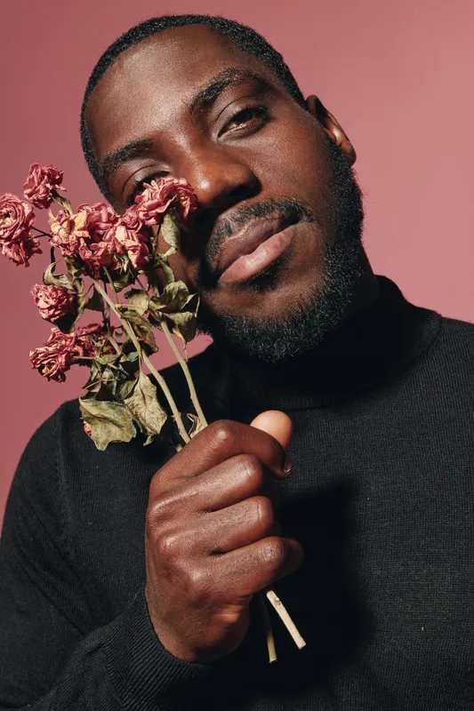 African man with dark skin holding dried flowers close to face in creative conceptual visual style. Portrait with pink background and black turtleneck sweater for artistic expression.