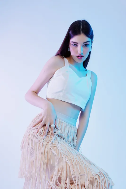 Young woman beauty fashion posing in a white crop top and fringed skirt with long dark hair. Studio shot with soft blue and pink lighting. Concept of style and elegance.