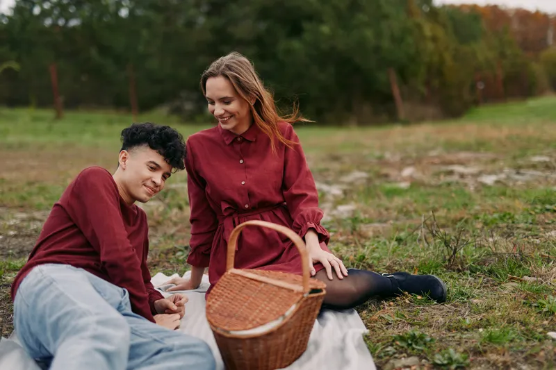 A candid outdoor picnic scene of a young couple sharing a quiet moment on a blanket, conveying authentic connection, natural smiles, relaxed posture and everyday warmth.
