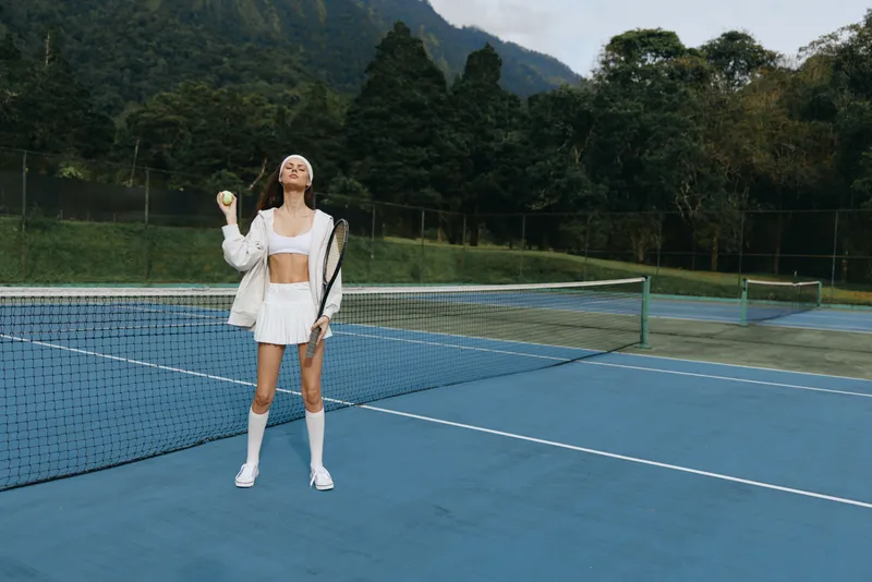 Young female tennis player celebrating on a court with mountains in the background, wearing a stylish white outfit, conveying joy and determination