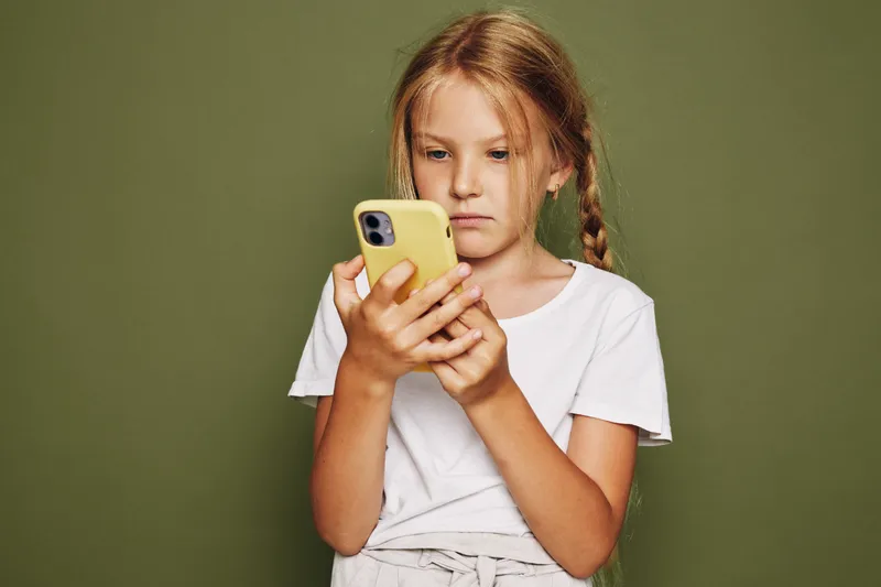 Young girl with braided hair wearing a white t-shirt concentrates on her smartphone. Neutral green background. Concept for childhood, technology, and communication.