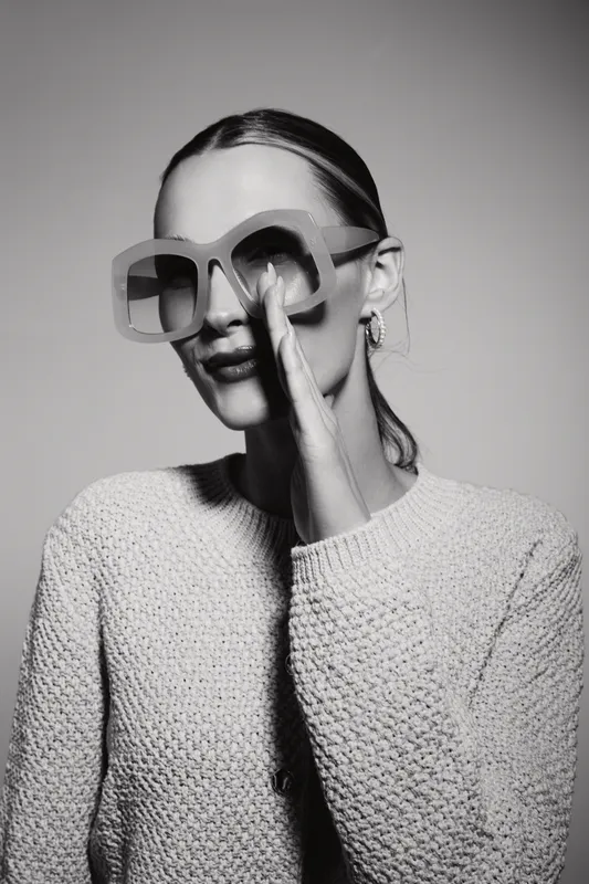 Stylish Caucasian woman with pale skin wearing oversized sunglasses and knitted sweater. She smiles subtly and touches her face, posing in a monochrome studio setting.