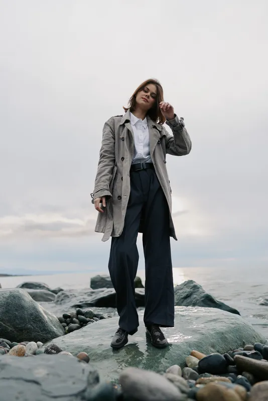 Young woman standing on rocky shore wearing a long coat and dark pants under overcast sky. Casual fashion concept with moody natural light and serene ocean background.