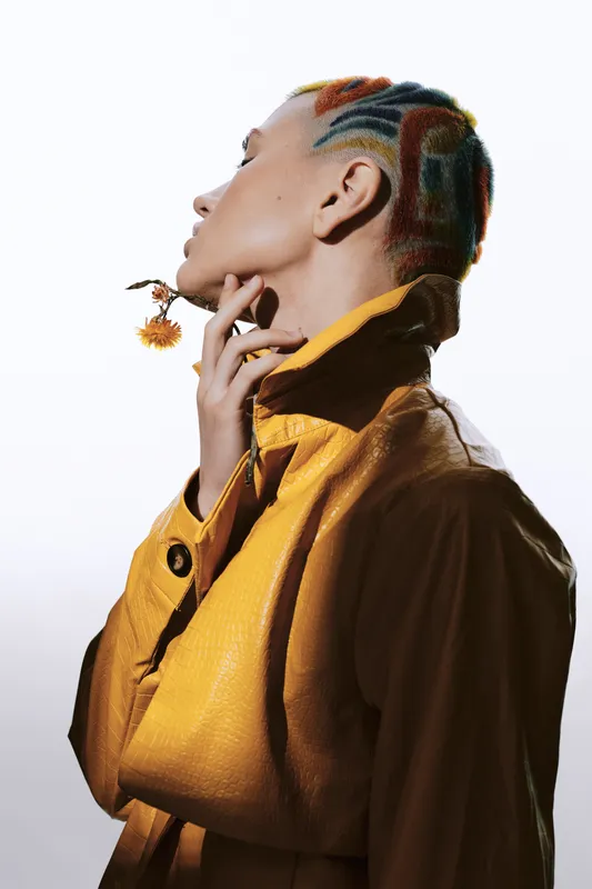 Woman with colorful braided hair and yellow jacket holds a flower, showcasing artistic style and unique self-expression against a clean white background.