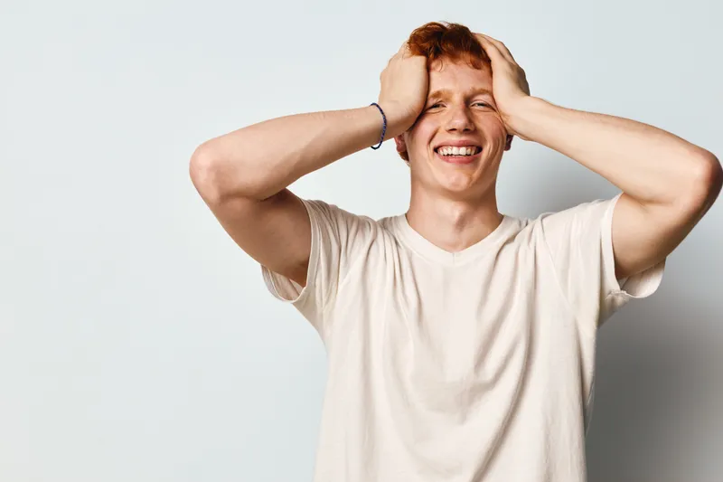 Young man with red hair laughing holding head with both hands against white background. Casual smiling male person with happy emotion and relaxed pose.