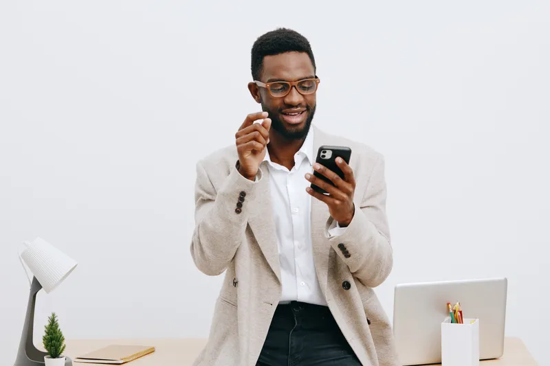 Smiling young man with glasses using smartphone in modern office environment, wearing beige blazer and white shirt, standing at his desk with laptop and stationery, looking happy and engaged, white ba