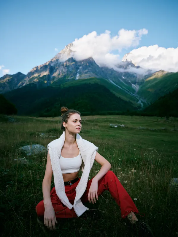 Outdoor scene with a woman seated in a grassy meadow, mountains above and clouds in a clear blue sky, casual athletic wear, relaxed pose, nature and solitude in natural light and open space