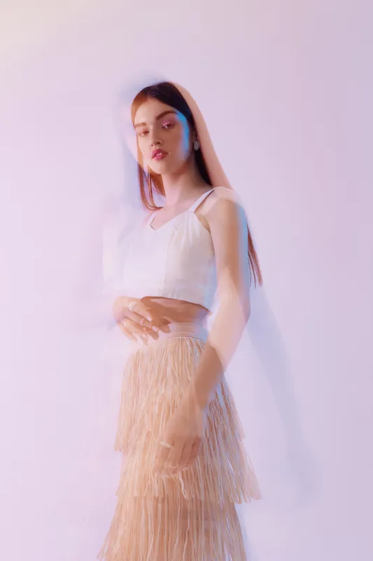 Young woman in a white top and beige fringed skirt poses against a light background. Soft lighting highlights her beauty and fashion style in a studio setting.