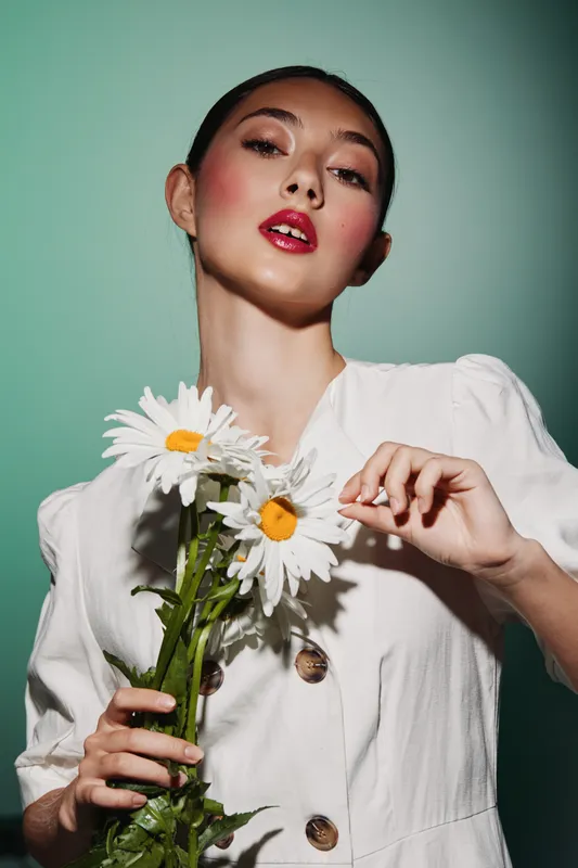 Young woman with bold red lipstick and smooth skin holds fresh white daisies against a green background. Fashion and beauty concept with cake creative shoot styling.