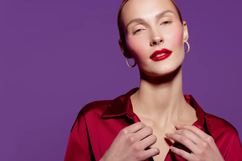 Portrait of confident woman with red lipstick and hoop earrings wearing red satin blouse on purple background. Concept of fashion, beauty, elegance, and modern style for editorial or advertising.