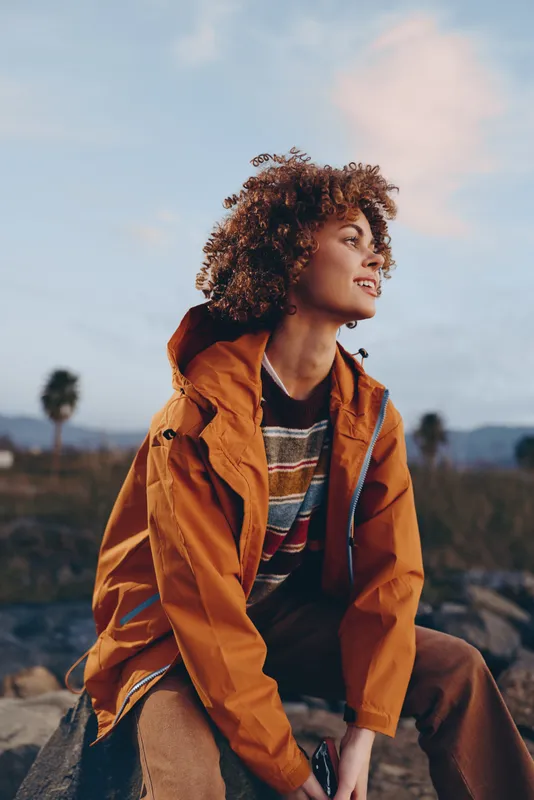 Outdoors lifestyle portrait of a woman with curly hair in an orange jacket, sitting at sunset on a hill, calm smile, soft light, distant mountains behind, travel mood.