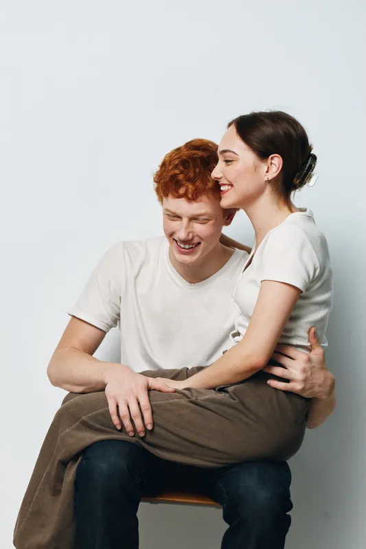 Couple, love, happiness, smile and relationship with woman sitting on man’s lap for affection, tenderness and joy. Young people, casual clothes and emotional connection in studio with white