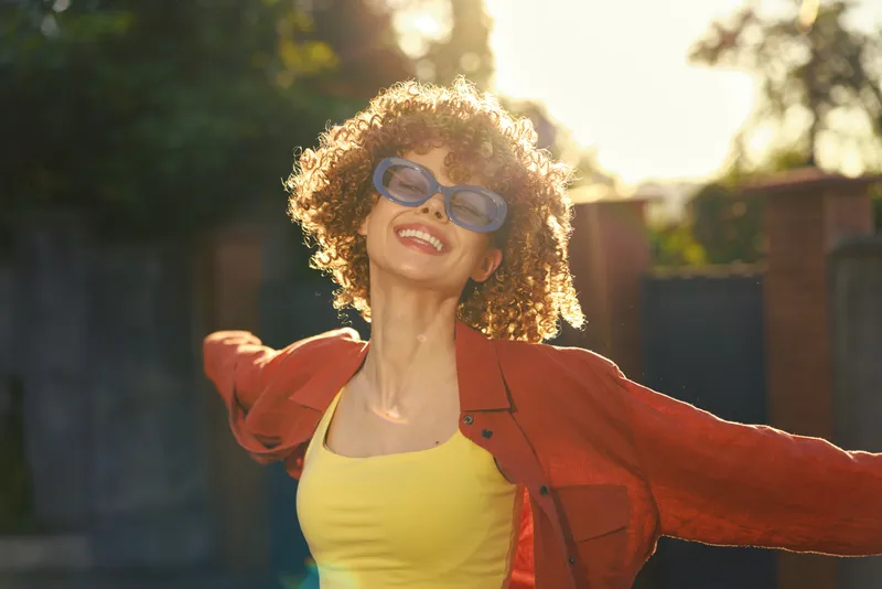 Joyful woman outdoors, wearing a yellow top and red jacket, with blue sunglasses, arms outstretched, sunlit moment, candid smile, breeze in hair, vibrant colors, carefree lifestyle