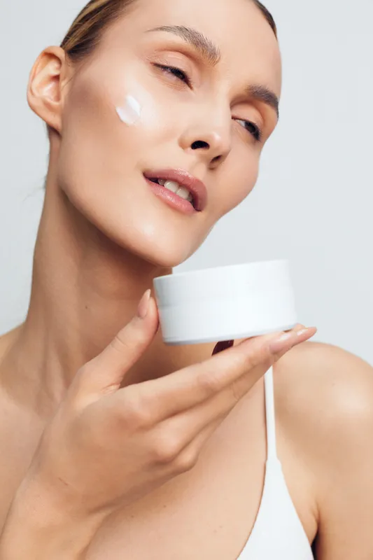 A young woman applies cream to her face in a clean studio, capturing a focused skincare moment that conveys self-care and calm, with a soft glow on her skin.