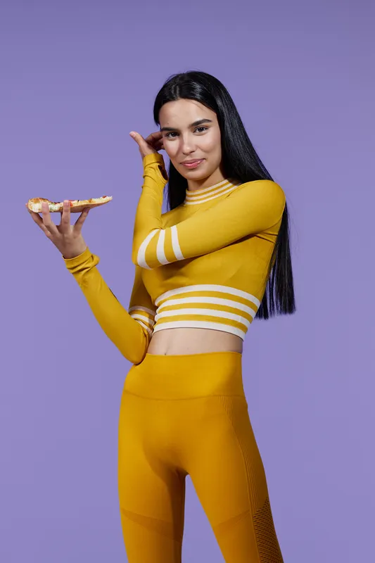 Woman sport fitness wearing yellow activewear holding a slice of pizza, slender and healthy, standing against a purple background, casual and energetic mood.