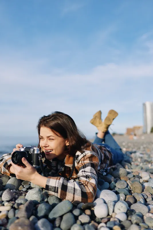 Woman photographer lying on a rocky beach with a camera, capturing candid outdoor moments. Casual plaid shirt, relaxed pose, sea breeze and clear sky frame a serene coastal scene of nature photography