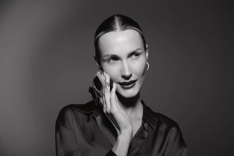 Caucasian woman with pale skin talks on phone, wearing black silk blouse and hoop earrings, studio portrait in black and white.