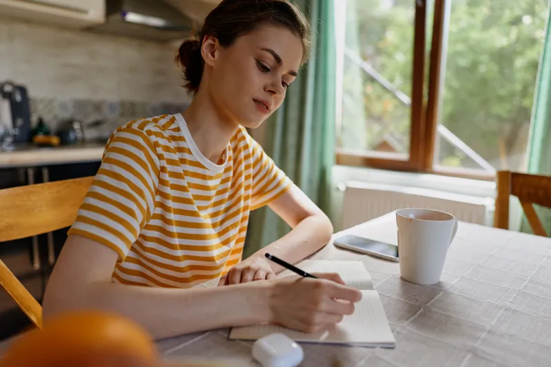 Writer woman writes in a notebook at a wooden kitchen table bathed in warm daylight by a window, capturing a calm home moment.