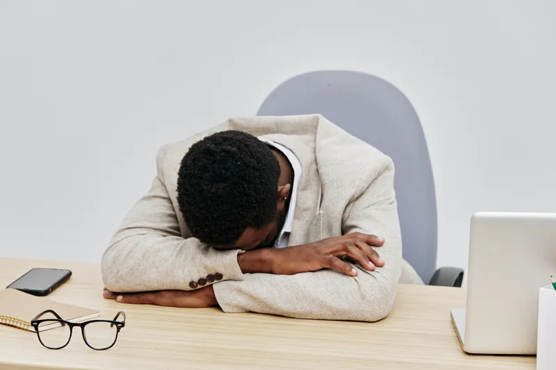 Overworked African American man resting his head on desk, feeling tired and exhausted after long working hours, workplace stress concept, indoors, casual beige blazer, modern office environment, subdu