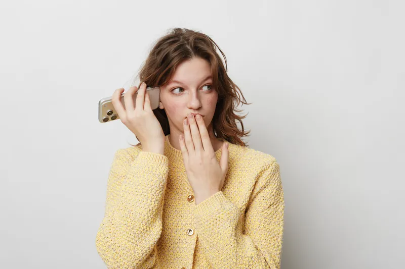 Young woman in yellow sweater holds smartphone near ear and covers mouth with hand, looking surprised. Concept of secret conversation, gossip, and unexpected news.
